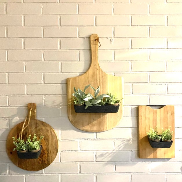 Three wooden cutting boards of different sizes with artificial plants in black pots attached, hanging on a white brick wall./DIY