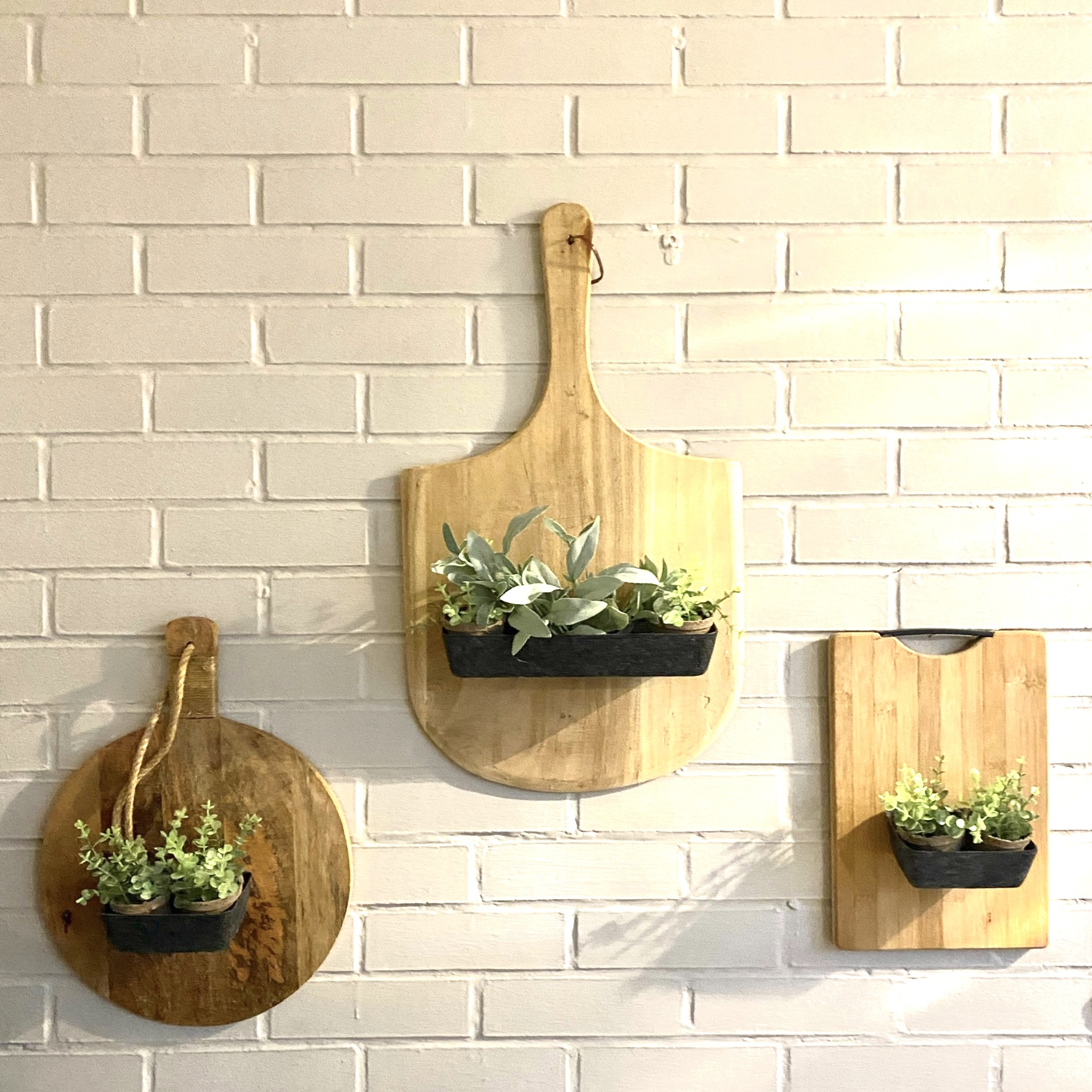 Three wooden cutting boards of different sizes with artificial plants in black pots attached, hanging on a white brick wall.