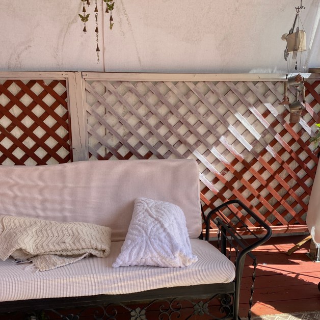 Outdoor patio area with a striped beige and white couch, decorative pillows, and a lattice backdrop under a shelter with hanging plant decorations and a lamp.