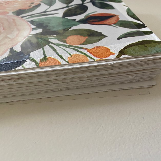 Stack of papers with visible floral and fruit watercolor designs resting on a white surface/creativity.