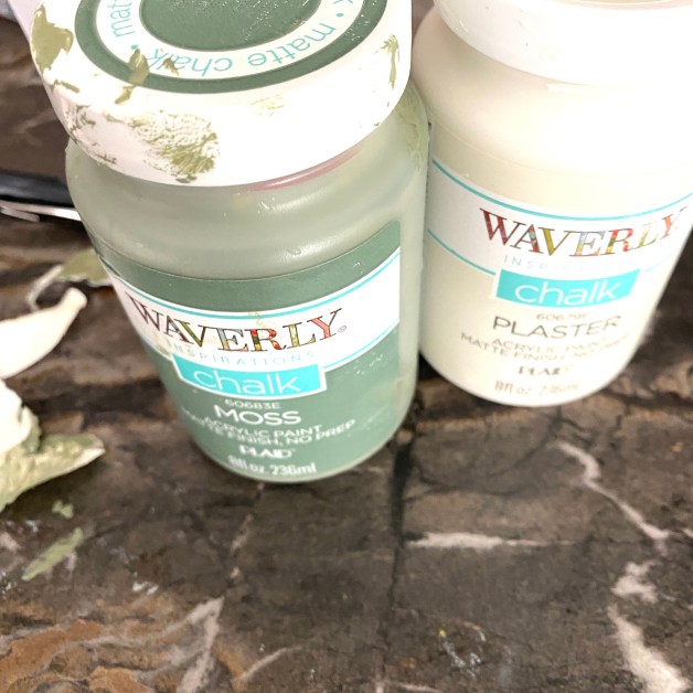 Two jars of Waverly Inspirations chalk paint in the colors 'Moss' and 'Plaster' on a marbled background.