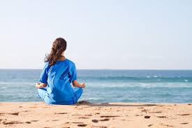 A person in a blue garment sitting in a meditative pose on a sandy beach, facing the ocean under a clear sky./words