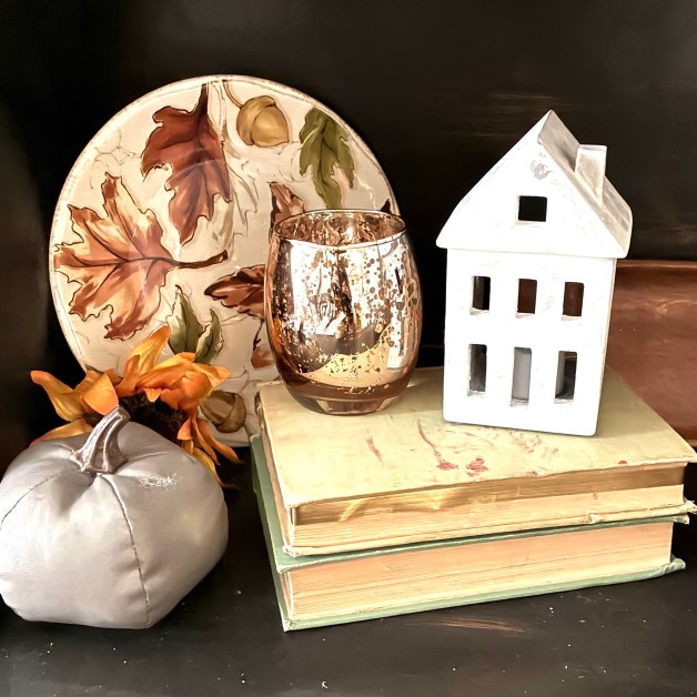 img_5922 | goodthoughtsgoodlives Autumn-themed still life: a leaf-patterned ceramic plate, a speckled glass goblet, a white decorative lantern shaped like a house, and fabric pumpkins on a shelf.