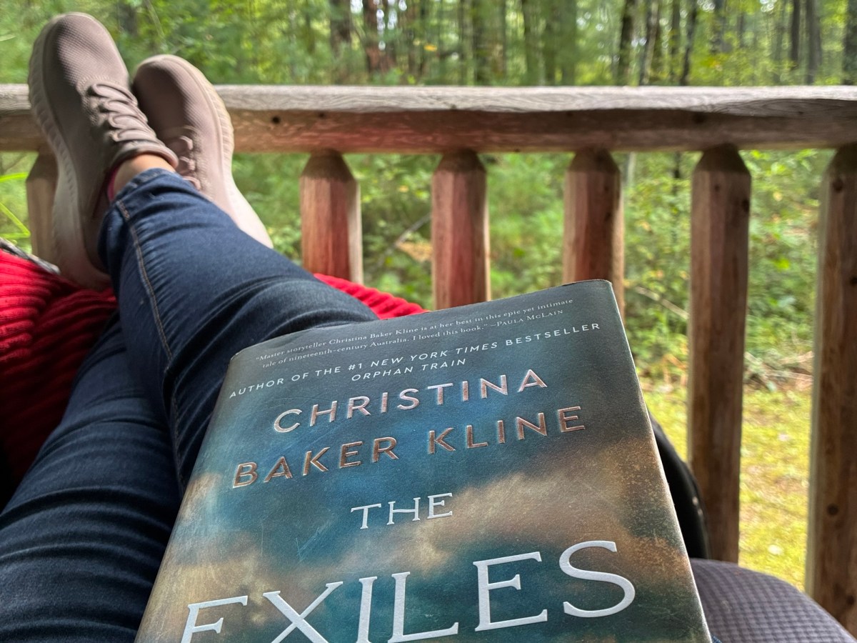 Person relaxing outdoors with a book on their lap, forest in the background and brown shoes visible on a wooden railing.