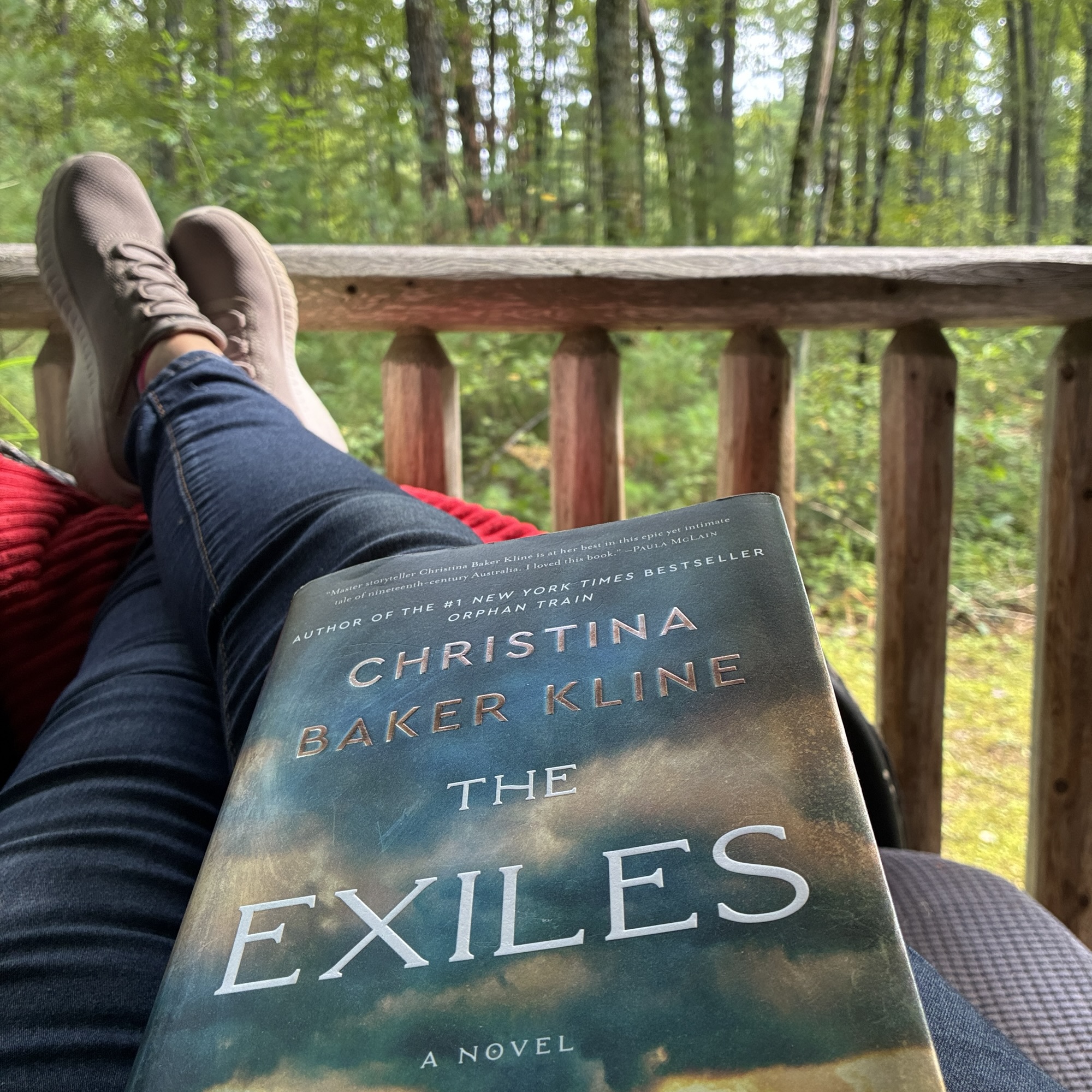 Person relaxing outdoors with a book on their lap, forest in the background and brown shoes visible on a wooden railing.