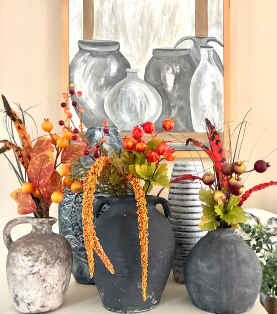 img_6020-1 | goodthoughtsgoodlives Four rustic ceramic vases with orange bead garlands and autumn foliage arranged in front of a grayscale painting of jars on a shelf.