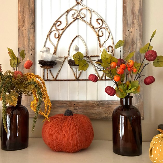 img_6024-1 | goodthoughtsgoodlives Fall mantel display with an orange pumpkin centerpiece, dried floral sprays, and two brown glass bottles against a white paneled backdrop.