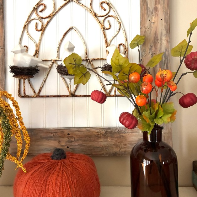 img_6026-1 | goodthoughtsgoodlives Fall harvest display featuring a large orange yarn pumpkin, orange berries on branches, and a rustic metal scrollwork behind a white paneled wall in a vase on the right side of the scene, creating a warm seasonal vignette.