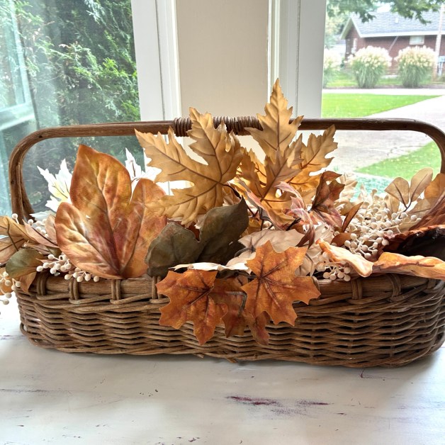 img_6028-1 | goodthoughtsgoodlives Wicker basket overflowing with dried autumn leaves in front of a sunlit window with a garden outside.